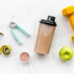 Fitness background with meal replacement shake, shaker, apple, dumbbells and measuring tape on stone table