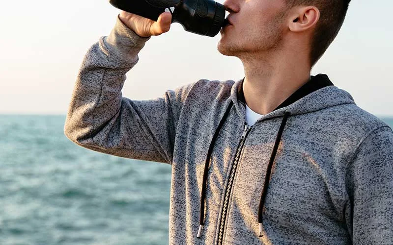 Sportsman drinking a water enhancer from bottle after running, workout, on quay, near the sea.