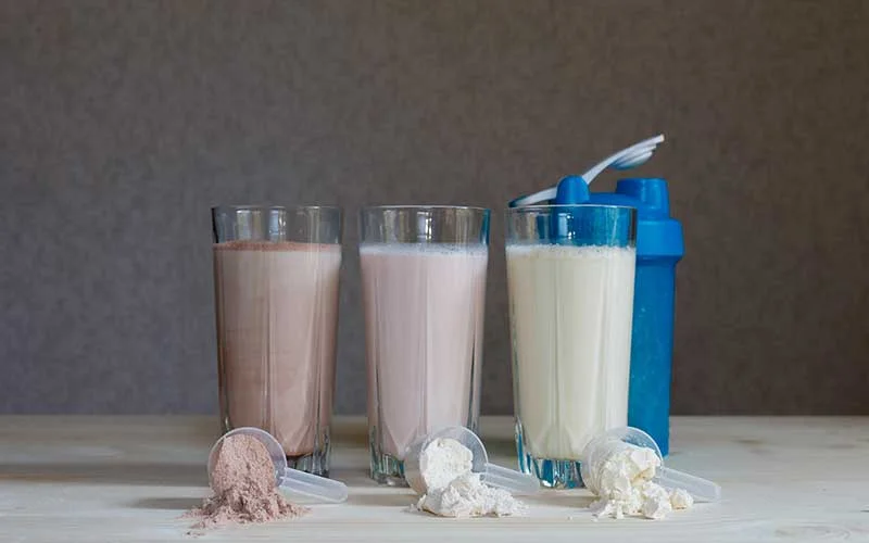 Three milk whey meal replacement shakes in glass cups are arranged in a row with three scoops and a blue shaker on a wooden table.