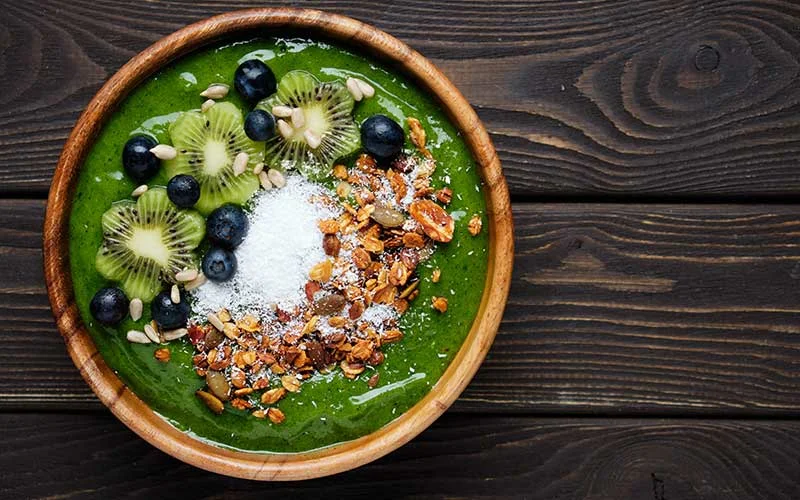 Green smoothie bowl with kiwi, blueberry, granola and coconut flakes on dark wooden background.