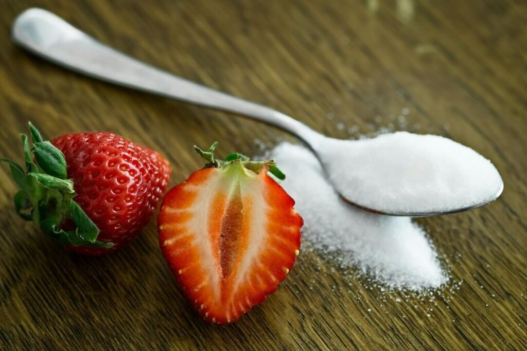 Fresh strawberries next to a spoonful of sugar on a table.