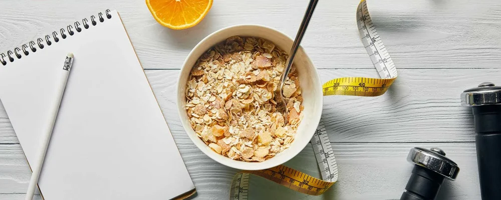 measuring tape, spoon and breakfast cereal in bowl near apple, orange, notebook, dumbbells and pencil on wooden white