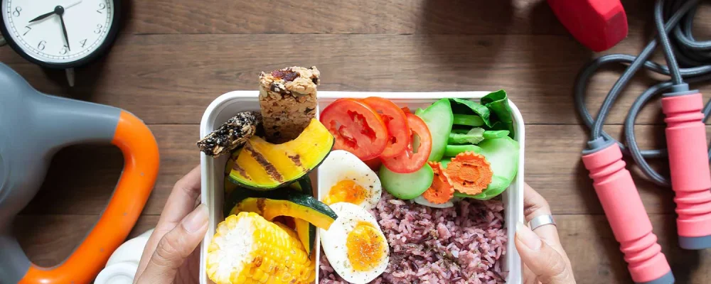 Woman's hands holding lunch box with rice berry, boiled eggs, sweetcorn, pumpkin, tomatoes and cereal bars, Top view with sport and fitness equipments