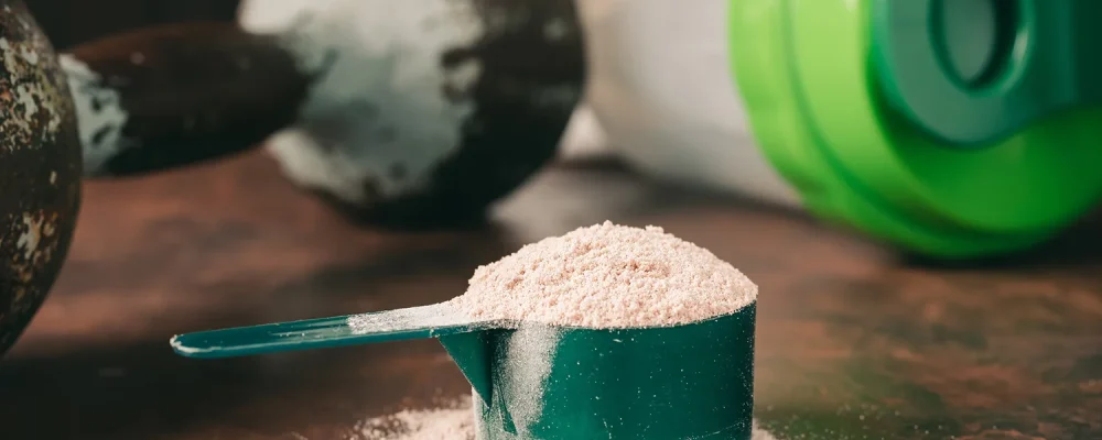 Chocolate meal replacement shake powder in measuring spoon, old rusty dumbbell and shaker on dark background