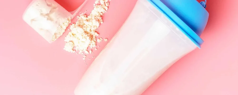 shaker and measuring spoon with meal replacement shake powder on a pink background